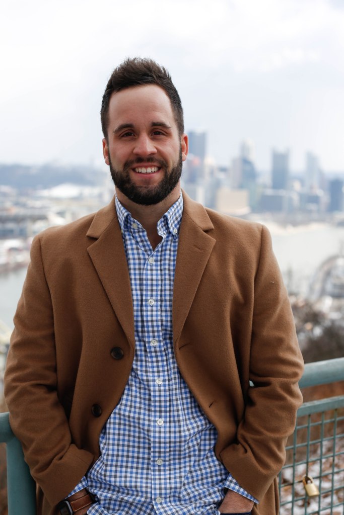 man smiling with blue and white checkered button up shirt and tan coat in front of city backdrop